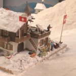 Miniature Swiss chalet in snowy Winter Mountain exhibit at Small Worlds Dubai with skiers, flags, and snow-covered scenery.