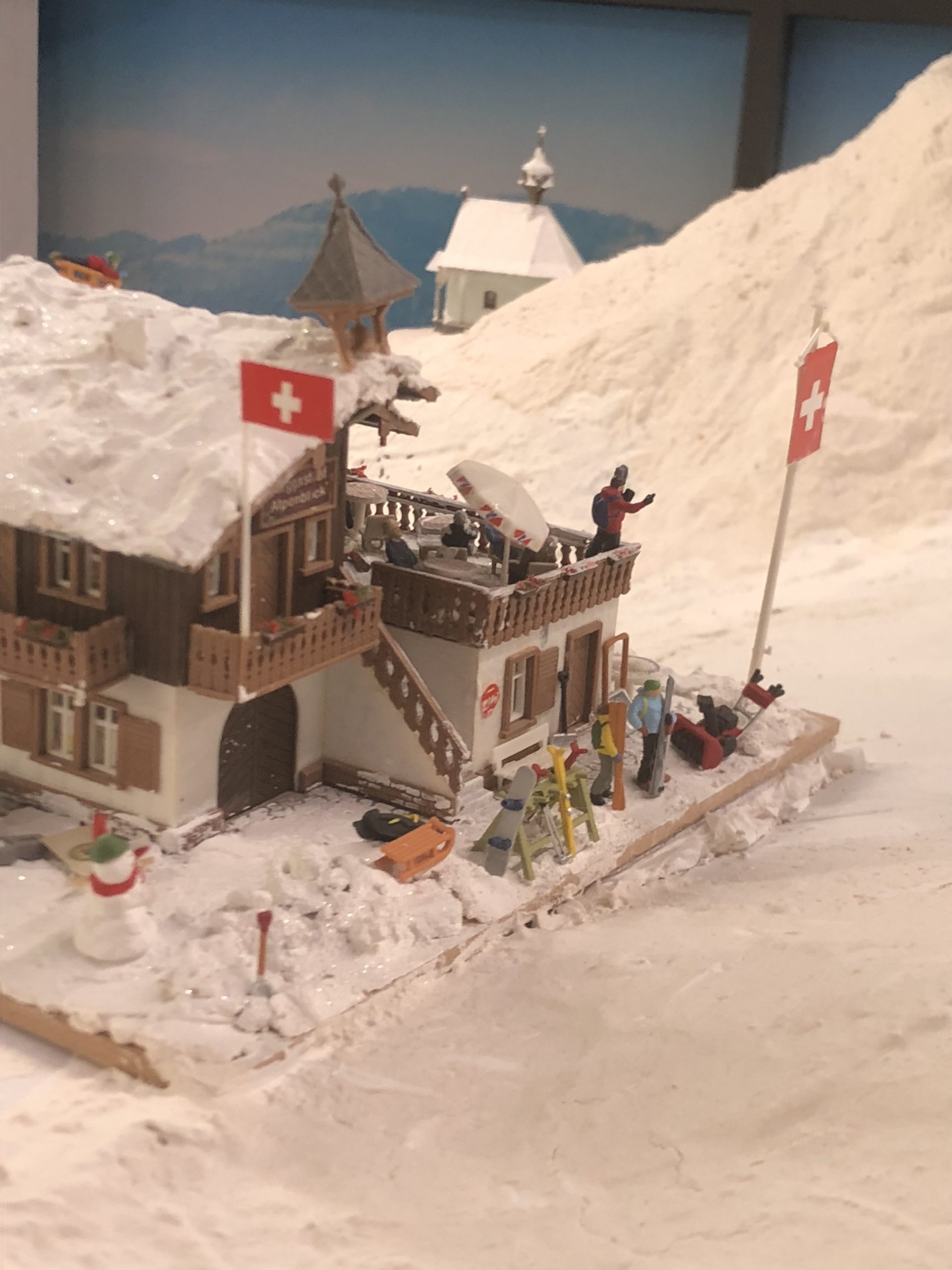 Miniature Swiss chalet in snowy Winter Mountain exhibit at Small Worlds Dubai with skiers, flags, and snow-covered scenery.